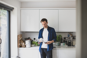 Adult male checking smartphone in kitchen with mug of coffee