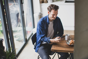 High angle shot of adult male checking smartphone in kitchen with mug of coffee