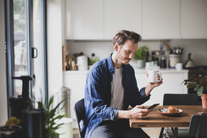 Adult male checking smartphone in kitchen with mug of coffee