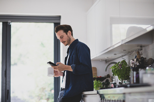 Adult male checking smartphone in kitchen with mug of coffee