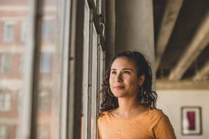 Young hispanic female looking out of window in loft apartment thinking