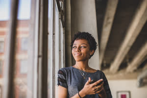 Young african american female looking out of window in loft apartment holding smartphone